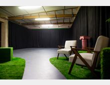 Ein minimalistischer Innenraum mit schwarzem Vorhang im Hintergrund, einer grauen Bodenfläche und künstlichem Rasen, der teilweise den Boden bedeckt. Im Vordergrund stehen zwei weiße Sessel mit Holzrahmen und eine rosafarbene, verzierte Säule.