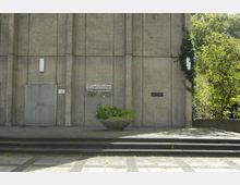 Außenansicht eines modernen Kirchengebäudes mit einer schlichten Betonfassade, einer Metalltür links und einem Schild mit der Aufschrift „Evangelische KFG Gemeinde“. Vor dem Gebäude stehen eine bepflanzte Betonschale und eine kleine Treppe, während rechts Pflanzen und Bäume zu sehen sind.