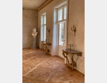 Eleganter Innenraum mit Fischgrät-Parkett, hohen weißen Fenstern und einer verglasten Tür, die viel Tageslicht hereinlässt. An den Wänden hängen vergoldete Wandkerzenhalter; dazu stehen zwei reich verzierte Konsoltische und eine Büste auf einer Säule.