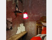 Ein rustikal eingerichtetes Schlafzimmer mit einer grob verputzten Wand, einer roten Wandlampe, zwei dekorativen Hundemotiven aus Holz auf einer Bank und einem goldfarbenen, verzierten Bettrahmen. Im Hintergrund sind Schwarz-Weiß-Collagen an der Wand angebracht.