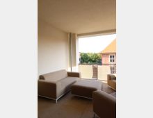 Überdachter Balkon mit beigefarbenen Polstermöbeln, bestehend aus zwei Sesseln, einem Sofa und einem Hocker. Im Hintergrund Glasgeländer und Blick auf grüne Bäume sowie ein Gebäude mit rotem Dach.