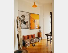 Flur in einer Wohnung mit hellem Holzfußboden und weißen Wänden. An der Wand hängen zwei große gerahmte Kunstwerke, darunter ein Schwarz-Weiß-Porträt und ein orangenes Bild; vor der Wand steht eine mit Kissen und einer Decke dekorierte Bank. Ein orangefarbener Deckenspot, eine Holzskulptur auf einem Tisch und moderne Dekorationsstücke ergänzen den Raum.