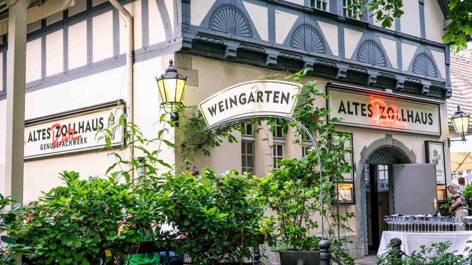 Fassade eines historischen Fachwerkhauses mit den Schildern „Weingarten“ und „Altes Zollhaus“, mit Laternen an der Wand und einem Rundbogen-Eingang. Davor stehen üppige Grünpflanzen in Kübeln und ein kleiner Außenbereich mit Tischen im Hof.