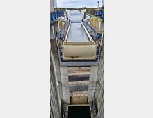 Moderne Schiffshebewerkanlage mit vertikalen Betonwänden, Wasserbecken und blauen Geländern. Im Hintergrund ist ein Wasserweg zu sehen, der von Bäumen und Wegen flankiert wird.