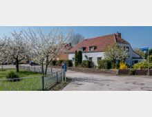 Wohnsiedlung mit einem weißen Haus mit rotem Satteldach, umgeben von blühenden Bäumen, Blumenbeeten und Rasenflächen. Im Vordergrund führt ein Gehweg mit dekorativem Metallzaun entlang, während Straßenschilder auf Straßennamen hinweisen.