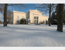 Klassizistisches Gebäude mit heller Fassade und Säulenportal, umgeben von verschneitem Gelände und Bäumen. Der Himmel ist blau, und das Gelände ist winterlich ruhig.