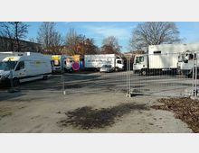 Ein eingezäunter Parkplatz mit mehreren geparkten Lieferwagen und Lastwagen, darunter Fahrzeuge mit der Beschriftung "MAIER BROS." und "Sixt". Im Hintergrund sind Bäume ohne Laub und Gebäude zu sehen.