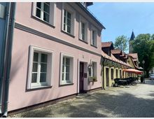 Eine gepflasterte Gasse mit zweigeschossigen historischen Gebäuden; links ein rosafarbenes Haus mit weißen Fensterrahmen, rechts gelbe Häuser mit roten Ziegeldächern und Außensitzplätzen mit Tischen und Sonnenschirmen. Im Hintergrund ist der Turm einer Kirche erkennbar.