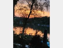 Sonnenuntergang an einem See mit einem hölzernen Steg im Vordergrund, umgeben von Bäumen und dichter Vegetation. Das Wasser spiegelt das orangefarbene Licht des Himmels, und im Hintergrund sind Boote am Ufer zu erkennen.