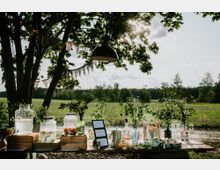 Ein rustikaler Bartisch im Freien steht unter schattigen Bäumen, dekoriert mit Gläsern, Getränkespendern und frischen Kräutern. Im Hintergrund ist eine grüne Wiese mit Bäumen und hellem Himmel sichtbar, während Wimpelketten an den Ästen hängen.