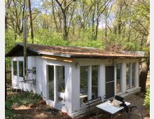 Kleines, weißes Gartenhaus mit Flachdach, teilweise mit Laub bedeckt, umgeben von einem dichten Wald. Das Haus hat große Fenster und eine Glastür, davor steht ein Tisch mit Stühlen auf einem gepflasterten Bereich.