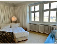 Ein modernes Schlafzimmer mit einem großen, weißen Bett vor bodenlangen cremefarbenen Vorhängen. Rechts im Raum befinden sich hohe Fenster mit Blick auf eine Stadtlandschaft, darunter Heizkörper mit weißem Gitter. Links hängt eine runde Lampe, und auf einer Kommode rechts stehen mehrere Glasflaschen.