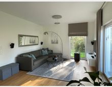 Modernes Wohnzimmer mit einer grauen Eck-Couch und mehreren blauen Kissen, einem runden schwarzen Couchtisch auf einem Teppich und einer Stehlampe mit gebogenem Arm. An einer Wand hängen ein großer Spiegel und ein Bild, gegenüber befindet sich ein Fernseher auf einem weißen Regal. Bodentiefe Fenster bieten Blick auf einen Garten.