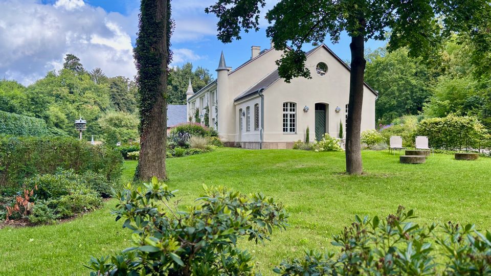 Helles, villaähnliches Gebäude mit Satteldach, Rundfenster und kleinen Türmchen, umgeben von einem gepflegten Garten mit Rasenflächen, Sträuchern und großen Bäumen. Im Vordergrund liegen Beete und Hecken, rechts stehen zwei schlichte Sitzgelegenheiten auf der Wiese.