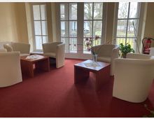 Ein gemütlicher Wartebereich mit cremefarbenen Sesseln und niedrigen Holztischen auf einem roten Teppichboden. Im Hintergrund sind große Fenster mit Blick auf eine grüne Außenanlage, und dekorative Pflanzen sowie ein Feuerlöscher ergänzen den Raum.