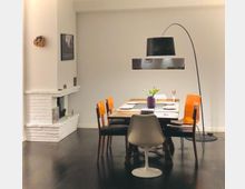 Esszimmer mit modernem Design: Ein Holztisch mit fünf verschiedenen Stühlen, darunter ein weißer, steht auf einem dunklen Boden. Im Raum befindet sich ein weiß gestrichener Kamin, eine Bogenlampe mit schwarzem Lampenschirm und eine Vase mit Tulpen auf dem Tisch.