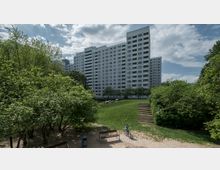 Hochhaus in einer Wohnsiedlung mit weißer Fassade, im Vordergrund ein Park mit Bäumen, Rasenflächen, einem Treppenweg und einer Person auf einem Fahrrad. Der Himmel ist leicht bewölkt.