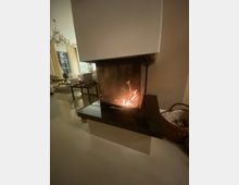 Ein Wohnzimmer mit einem modernen, eingebauten Kamin in einer Ecke, in dem ein Feuer brennt. Im Hintergrund sind eine helle Sitzgruppe, bodenlange Vorhänge und ein Kronleuchter sichtbar.