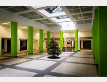 Offener Eingangsbereich eines modernen Gebäudes mit grünen Säulen, gefliestem Boden und einem gläsernen Deckenabschnitt, der Tageslicht hereinlässt. Ein geschmückter Weihnachtsbaum steht zentral, umgeben von Bänken und einem Informationsschalter im Hintergrund.