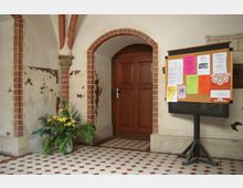 Kircheninnenraum mit einem hölzernen Pfeilertürrahmen und einer geschlossenen Tür. Links steht ein Blumenarrangement mit gelben Blüten, rechts eine Pinnwand mit bunten Aushängen; der Boden ist mit rot-weißem Fliesenmuster gestaltet.