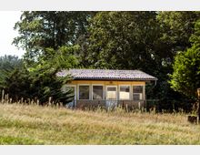 Ein einstöckiges Gebäude mit gelber Fassade und großen Fenstern, umgeben von hohen Bäumen und dichter Vegetation, liegt inmitten einer grasbewachsenen Fläche. Das Dach ist mit dunklen Dachziegeln gedeckt.