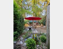 Ein Garten mit einem kleinen Teich im Vordergrund, umgeben von Steinen und Pflanzen. Im Hintergrund stehen Holztische und -stühle auf einem gepflasterten Bereich, beschattet von einem roten Sonnenschirm und hohen Bäumen.