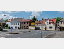 Straßenkreuzung in einem Wohngebiet mit kleinen, zweigeschossigen Häusern, teils verputzt und teils aus Ziegelstein, mit roten Dachziegeln. Verkehrsschilder wie Vorfahrts- und Richtungsschilder stehen an den Straßenecken, umgeben von einer ruhigen, ländlichen Umgebung bei sonnigem Wetter.