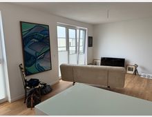Wohnzimmer mit hellen Wänden, Holzboden und großen Doppeltüren mit Blick auf einen Balkon. Im Raum stehen ein beiger Sofa, ein Flachbildfernseher, mehrere Bilderrahmen und eine moderne Lampe; an der linken Wand hängt ein abstraktes Gemälde.