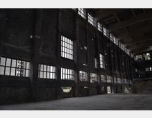 Innenansicht einer verlassenen Fabrikhalle mit hohen, dunklen Wänden und mehreren großen, rechteckigen Fenstern, die brüchige Glasflächen zeigen. Der Betonboden und die teilweise beschädigte Fassade betonen den verfallenen Zustand des Gebäudes.