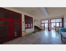 Flur eines Gebäudes mit roten Tür- und Fensterrahmen, gefliesten gelben Wänden und einer Treppe, die nach oben führt. Im Hintergrund sind Räume mit großen Fenstern und Möblierung erkennbar.