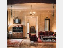 Wohnzimmer in einem historischen Gebäude mit abgenutzten Wänden und einem Holzboden im Fischgrätmuster. Es gibt rote Polstermöbel, einen Couchtisch aus Glas, einen Kronleuchter und einen Durchgang zu einem angrenzenden Raum mit antiken Möbeln.