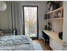 Schlafzimmer mit großem Fenster, das auf einen kleinen Balkon und Bäume hinausblickt. Links steht ein Bett mit heller Bettwäsche, rechts befindet sich ein Schreibtisch aus Holz mit Regalen und Schreibutensilien, darüber Pflanzen und Dekoration.