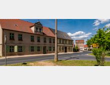 Straßenszene in einer ländlichen Ortschaft mit einem zweigeschossigen Gebäude mit roten Ziegeldächern und grünen Fensterläden im Vordergrund. Eine gepflasterte Straße führt um eine Kurve, flankiert von Bäumen und einem Verkehrsschild, im Hintergrund sind weitere historische Gebäude sichtbar.