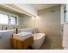 Modernes Badezimmer mit einem freistehenden weißen Waschbecken auf einem Holzunterschrank, einer ovalen Badewanne und einer großen Wandspiegel-Leuchte. Wände und Boden sind in neutralen Beigetönen gehalten, mit einem Fenster und einer Glasabtrennung im Hintergrund.