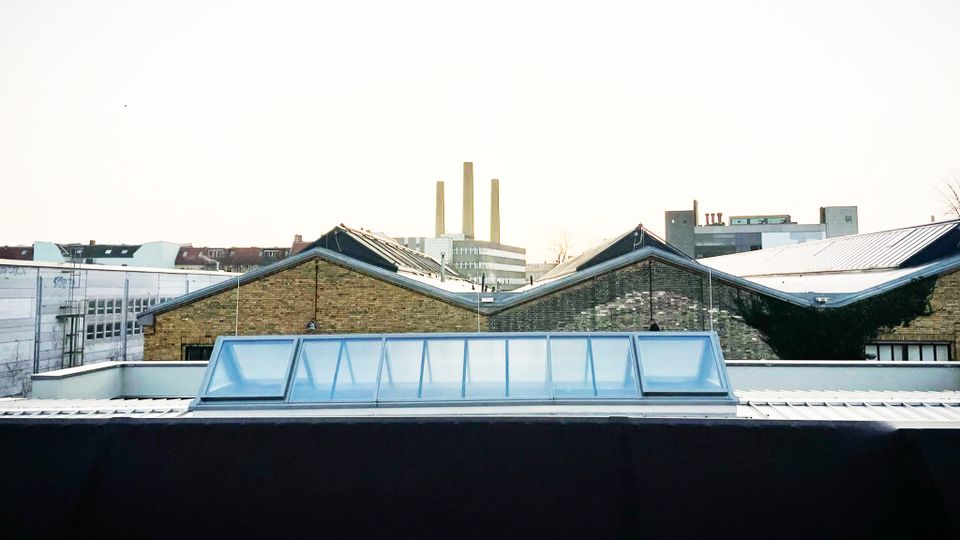 Gebäude mit gemauerten Fassaden und Satteldächern in einem städtischen Gebiet, im Vordergrund ein modernes Dach mit einer gläsernen Oberlichtkonstruktion, im Hintergrund industrielle Schornsteine und moderne Bauwerke.