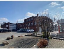 Gewerbegebiet mit einem großen Backsteingebäude im Hintergrund, das mehrere Rundbogenfenster hat, und eine Werbetafel mit der Aufschrift „Raab Karcher“. Im Vordergrund gepflasterte Flächen, einige Blumenbeete mit kargem Bewuchs und ein kahler Baum.