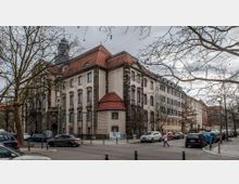 Straßenszene mit einem großen, historischen Gebäude aus hellem Putz und dunklem Stein, das ein rotes Ziegeldach hat. Der Himmel ist bewölkt, Bäume ohne Laub stehen entlang der Straße, und parkende sowie fahrende Autos sind sichtbar.