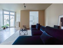 Wohnzimmer mit hellbeiger Wandfarbe, dunklem Holzboden und großer Glastür, die zu einem Balkon führt. Im Vordergrund steht ein dunkelblaues Sofa, ein runder Couchtisch mit Dekoration und im Hintergrund ist eine offene Verbindung zur weißen Küche sichtbar.