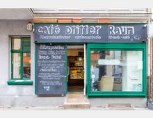 Außenansicht eines Cafés mit dem Namen "Café Dritter Raum". Die Fassade besteht aus einer schwarzen Tafel mit handgeschriebener Schrift, die Öffnungszeiten und Angebote wie Brunch beschreibt, sowie grünen Fenstern und einer Tür, die den Innenraum teilweise zeigt.