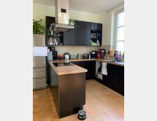 Moderne Küche mit dunklen Holzschränken, einer Kücheninsel mit integriertem Herd und einer Edelstahl-Dunstabzugshaube. Der Raum hat helle Wandfliesen, ein großes Fenster rechts und einen Holzboden im Fischgrätenmuster.