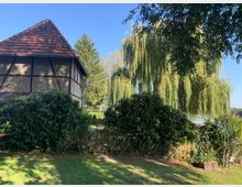 Fachwerkhaus mit rotem Ziegeldach am Rand eines Gartens, umgeben von grünen Hecken und Bäumen, darunter eine große Trauerweide, im Hintergrund ein See und blauer Himmel.