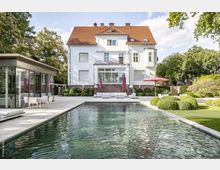 Großes weißes Wohnhaus mit rotem Ziegeldach und dekorativen Erkern, umgeben von gepflegtem Garten. Im Vordergrund befindet sich ein rechteckiger Pool, daneben eine moderne Glas-Pavillon-Struktur mit außenstehenden Möbeln und ein Sonnenschirm auf einer großzügigen Terrasse.