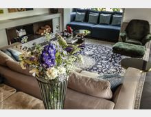 Ein elegantes Wohnzimmer mit einem großen Blumenstrauß in einer Glasvase im Vordergrund. Im Hintergrund befinden sich eine beige Couch, ein dunkler Teppich mit floralem Muster, eine dunkelblaue Couch mit Kissen und ein Sessel.