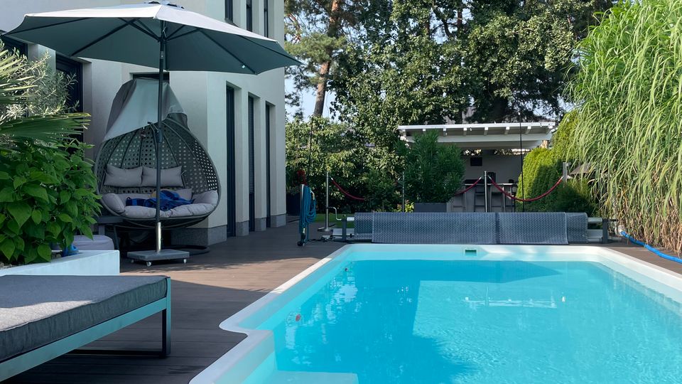 Modernes Wohnhaus mit hellem Putz und großen Fenstern, angrenzend an eine Terrasse mit Holzboden, einem rechteckigen Pool und Gartenmöbeln. Im Hintergrund befinden sich Bäume und eine kleine Überdachung, umgeben von dichter Vegetation.