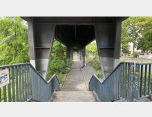 Ein schmaler Gehweg unter einer Stahlkonstruktion einer Hochbahn oder Brücke, flankiert von Geländern und umgeben von Bäumen sowie etwas städtischer Umgebung im Hintergrund. Das Metall der Konstruktion ist teilweise mit Graffiti besprüht.