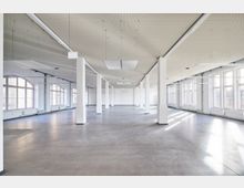 Weitläufiger, leerer Innenraum mit weißen Wänden und einer Reihe von rechteckigen Säulen. Der Raum hat große Fenster mit geschwungenen Oberlichtern, einen glatten Betonboden und eine perforierte Decke mit eingelassenen Leuchten.