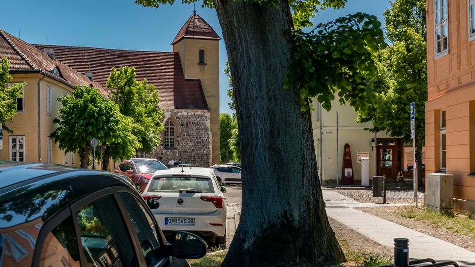 Straßenszene in einem historischen Ortskern mit geparkten Autos, großen Bäumen und niedrigen, hell verputzten Gebäuden mit Ziegeldächern. Im Hintergrund steht eine kleine Kirche mit steinernem Sockel und einem quadratischen Turm mit spitzem Dach.