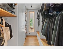 Schmaler begehbarer Kleiderschrank mit Holzfußboden, weißen Schränken und Regalen an den Seiten, sowie Kleiderstangen mit Kleidung. Im Hintergrund befindet sich ein schmales Fenster mit Blick auf Bäume und einen angrenzenden Balkon.