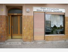 Fassade eines Gebäudes mit beige-braunen Kacheln, bestehend aus einer Holztür mit Hausnummer 28 und einem Schaufenster mit der Aufschrift „Freie evangelische Bibel-Baptisten-Gemeinde“. Im Fenster hängen weiße Gardinen und es sind Pflanzen zu sehen.
