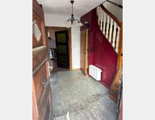 Eingangsbereich eines älteren Hauses mit abgenutztem Holzboden, einer roten Holzverkleidung an der Wand neben einer Holztreppe mit weißen Balustern. Im Hintergrund führen Türen in eine Küche und weitere Räume, an der Decke hängt eine mehrarmige Hängelampe.